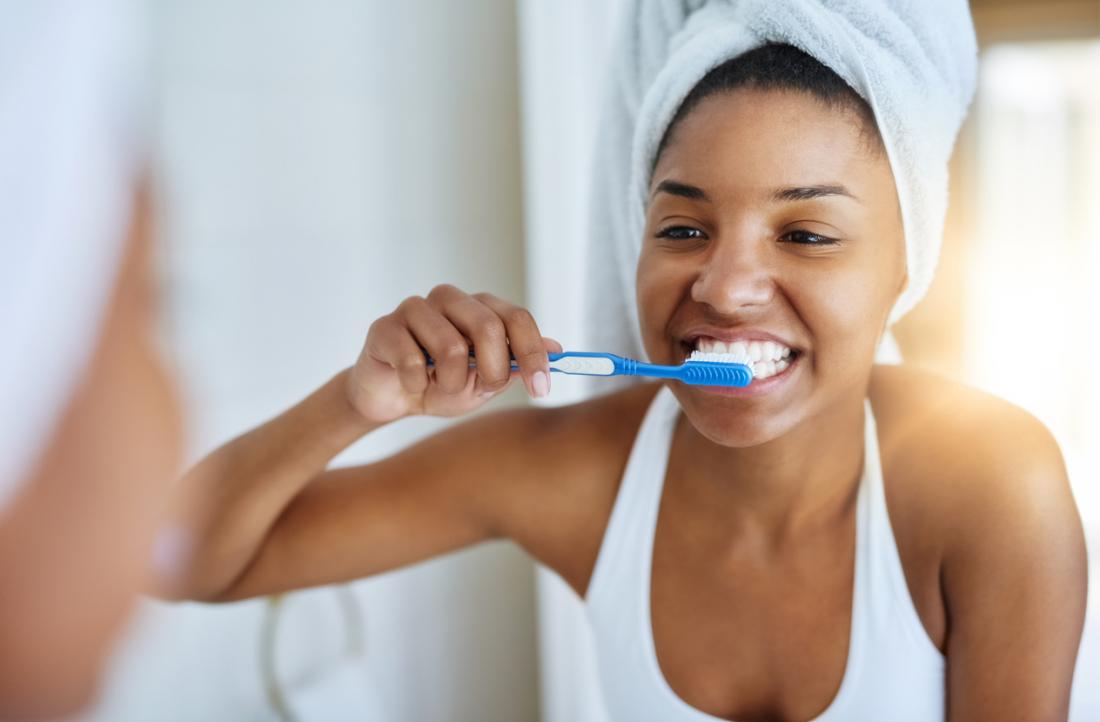 woman brushing her teeth in front of the bathroom mirror hair wrapped in a towel