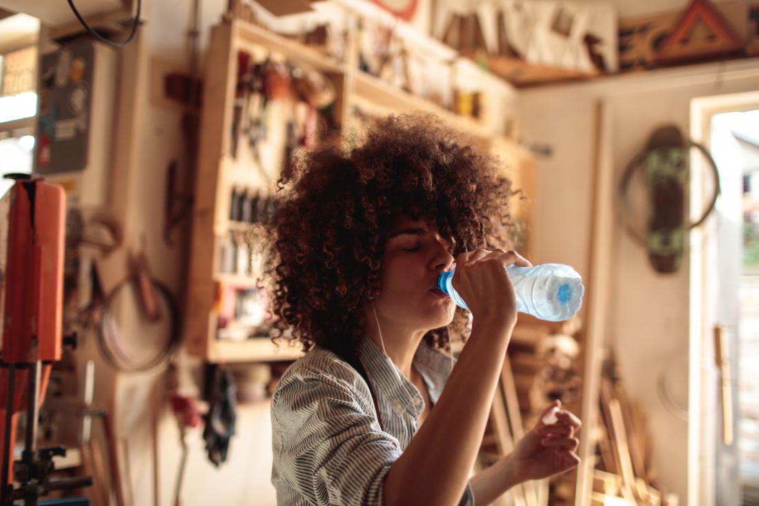 A woman who is thirsty after consuming baking soda drinks a bottle of water in a carpentry workshop.