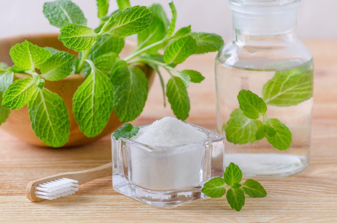 A wooden toothbrush, mint plant, glass pot of salt, and glass bottle of clear liquid, on a wooden table. Ingredients for natural salt mouthwash.