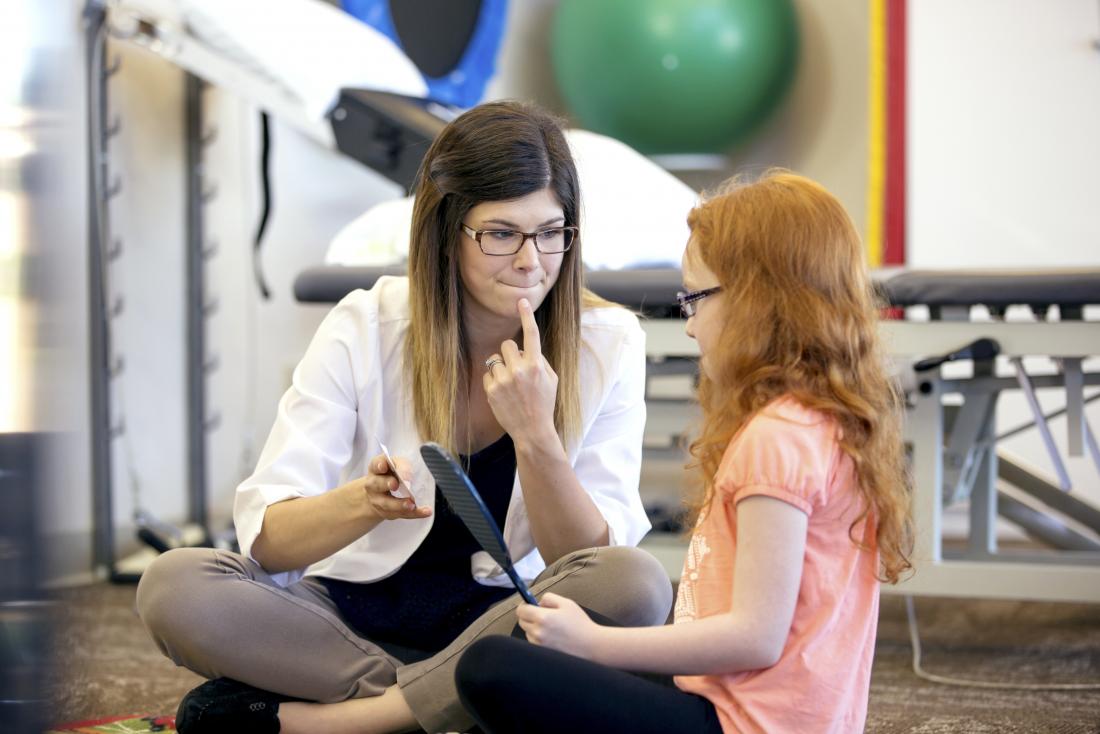 Speech therapy being conducted by doctor sitting with young girl, who is holding a mirror.