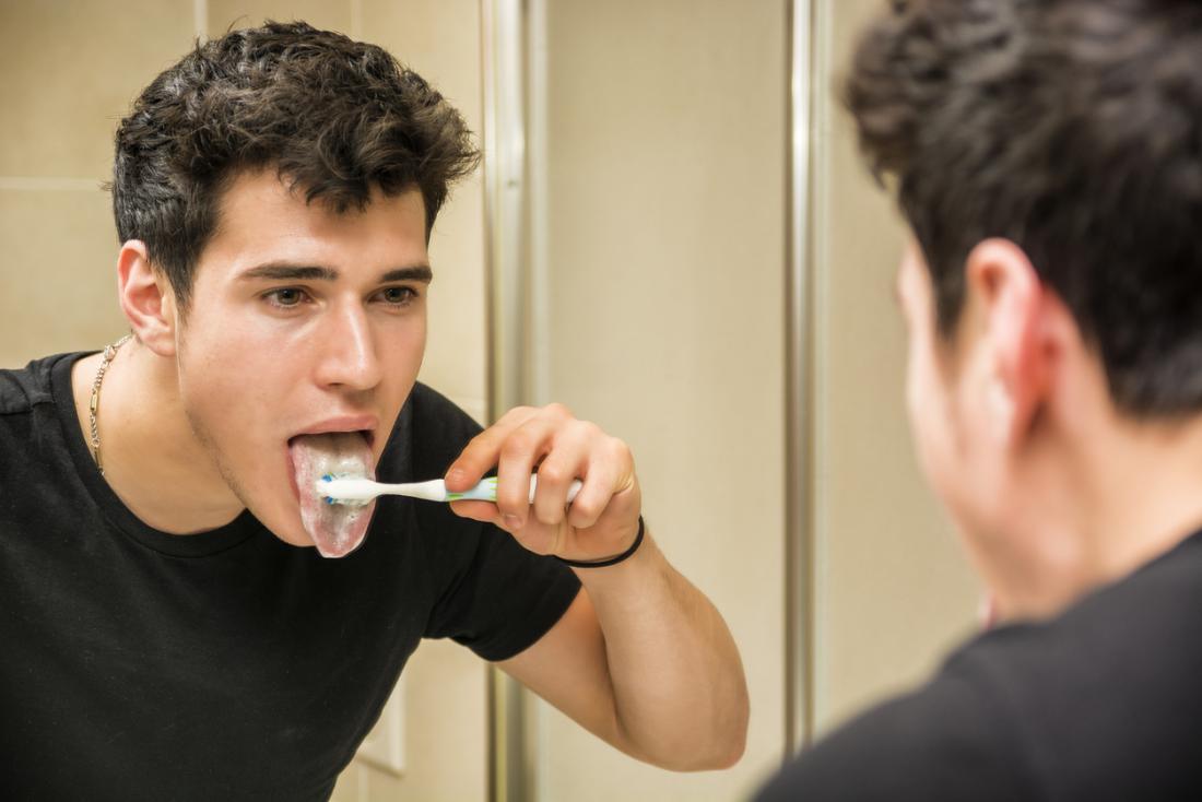 Young man brushing his tongue in the bathroom mirror